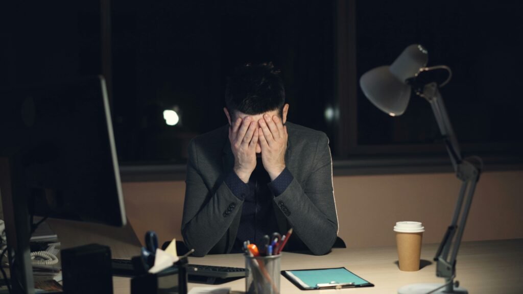 exhausted man at desk showing burnout symptoms, head in hands