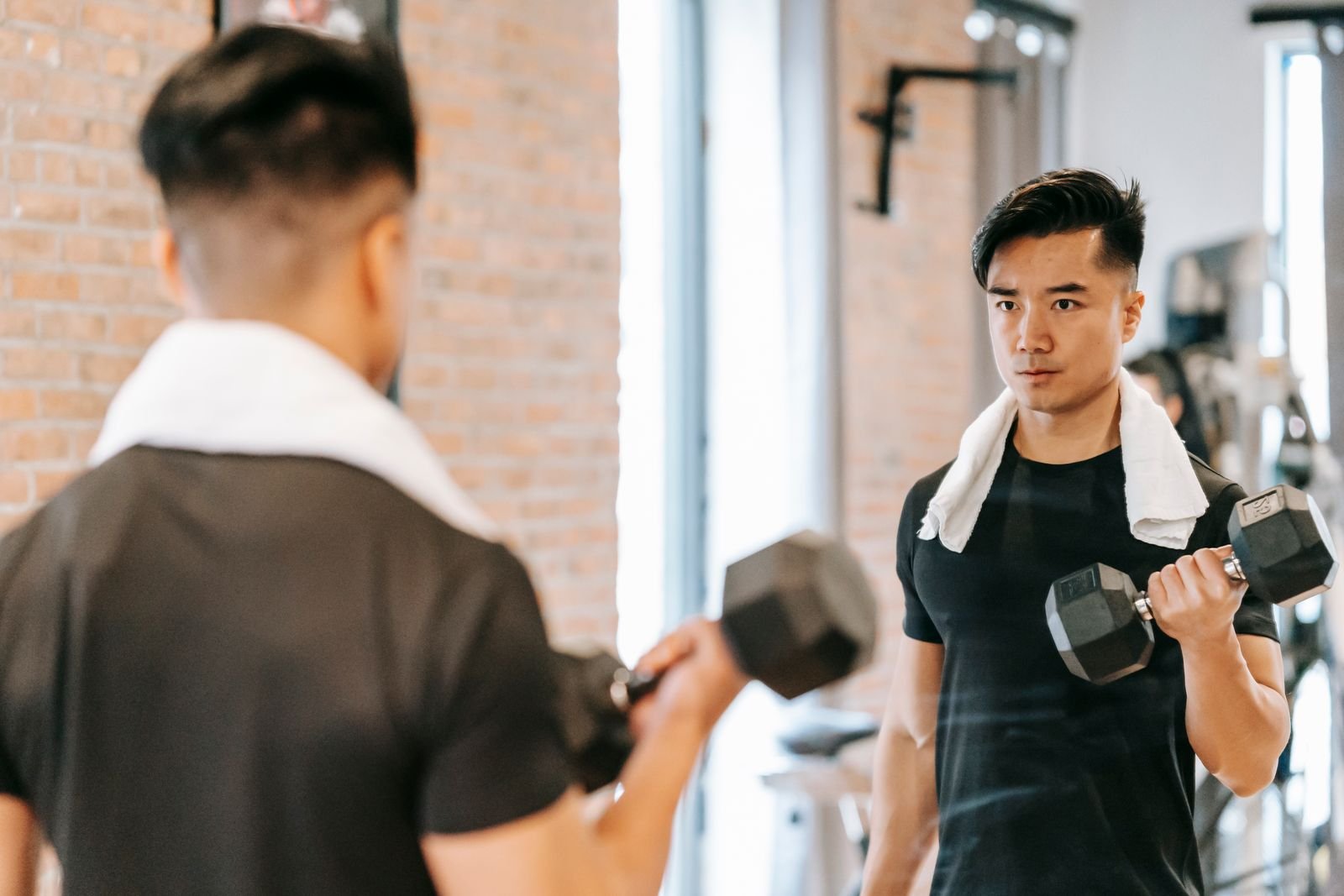 Man with a towel round his neck lifting a dumbbell in front of a gym mirror
